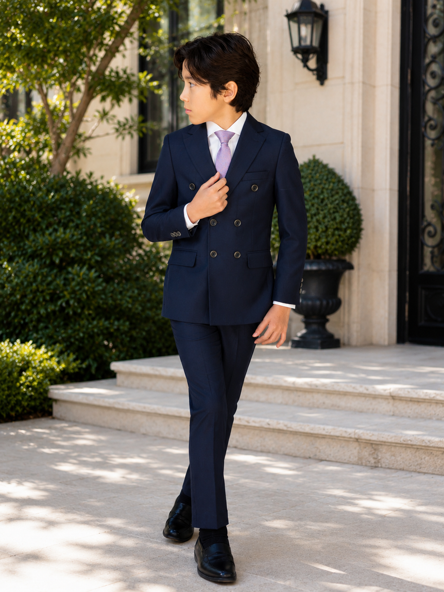 A young boy wearing a navy blue double-breasted suit, white dress shirt, and a light purple tie, standing on stone steps in front of a building.