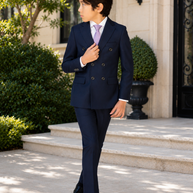 A young boy wearing a navy blue double-breasted suit, white dress shirt, and a light purple tie, standing on stone steps in front of a building.