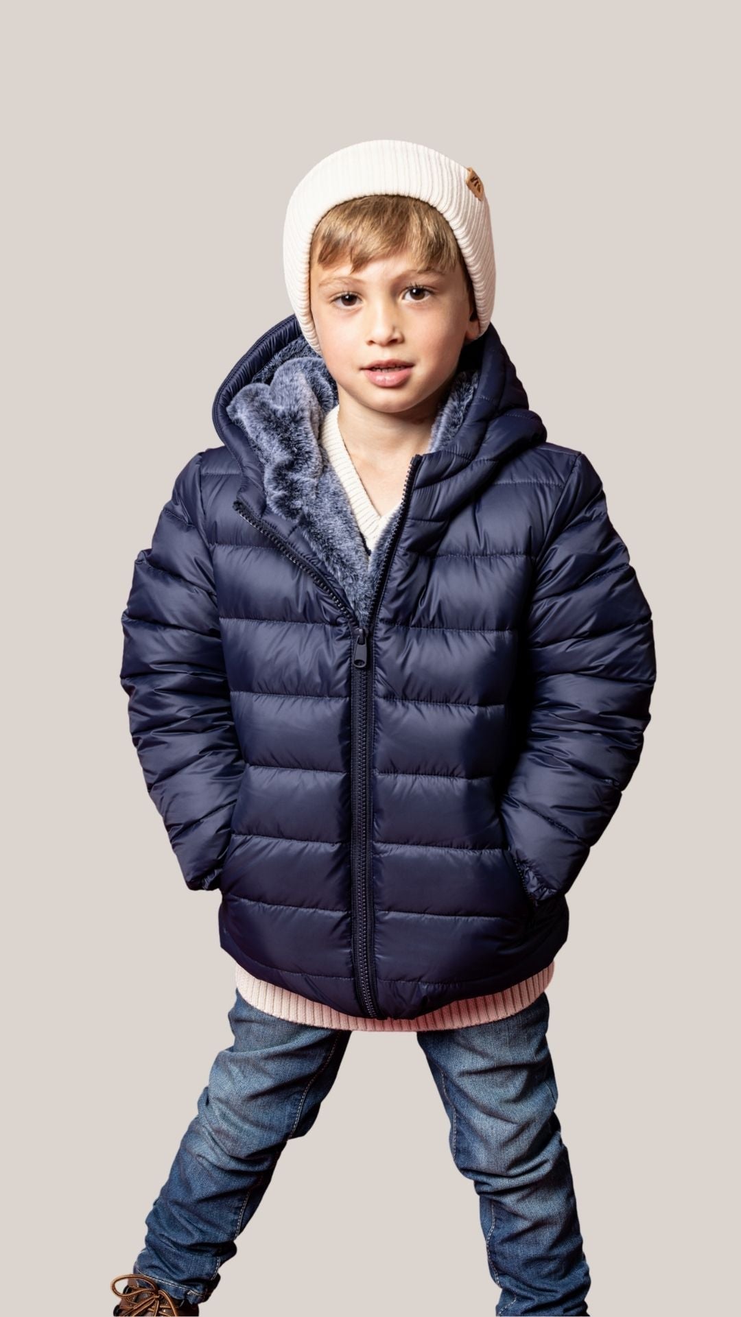 Child wearing a blue puffer jacket and white beanie on a beige background