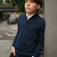 Young boy in a navy sweater and dark pants leaning against a wall outdoors.