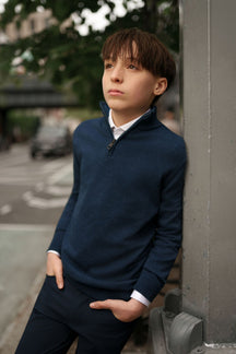 Young boy in a navy sweater and dark pants leaning against a wall outdoors.